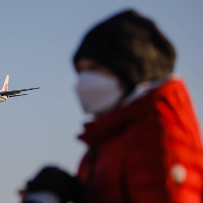 Ett flygplan avtecknar sig mot himlen. I förgrunden står en oskarp figur, men det är tydligt att personen bär munskydd, vintermössa och jacka.