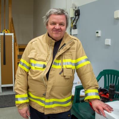 En medelålders man i gul brandmansjacka står inne i en hall och tittar leende in i kameran.
