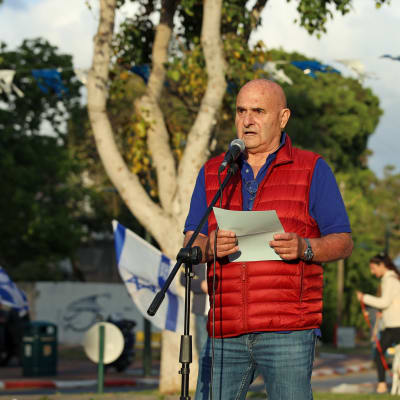 En äldre man i jeans och röd väst står på en scen och håller tal.