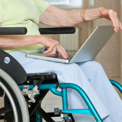 En äldre person sitter med bärbar dator i famnen i en rullstol.