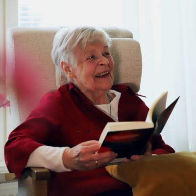 Iäkäs harmaahiuksinen nainen istuu nojatuolissa ikkunan edessä kirja kädessä ja hymyilee.