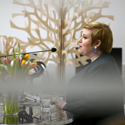 Annika Saarikko fotograferad från sidan medan hon står vid ett talarpodium och pratar. På talarpodiet finns en bukett tulpaner och ett glas vatten. Framför henne mikrofoner.