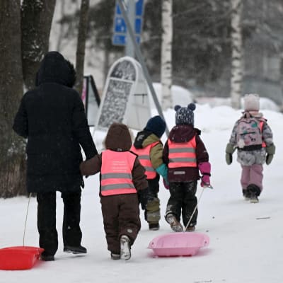 Lapsia kävelee jonossa lumisessa maisemassa yhen aikuisen kanssa. Aikuinen ja yksi lapsista vetää pulkkaa. Lapsilla on huomioliivit päällä.