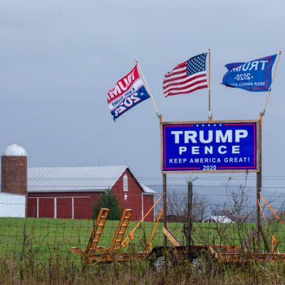 En skylt med texten Trump Pence Keep America Great 2020 på en äng med en lada i bakgrunden. På skylten har någon fäst tre flaggor: en amerikansk flagga och två olika Trump 2020-flaggor.
