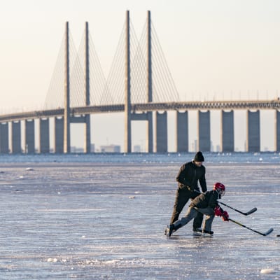 Man och barn spelar hockey på havsisen, med Öresundsbron i bakgrunden. 