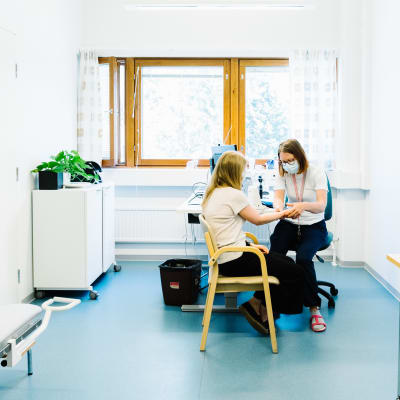 En hälsovårdare sitter med en patient i sitt rum. Hon håller patientens hand, det verkar som om hon undersöker något.