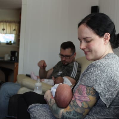 Kvinna som sitter i soffan med ett spädbarn i famnen, i bakgrunden sitter en man med ett annat spädbarn i famnen. 
