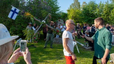 En reporterintervjuar en person, i bakgrunden reses en midsommarstång.