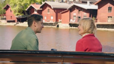 Två personer sitter vid ån och ser på varandra, i bakgrunden Borgås röda gammaldags byggnader. 