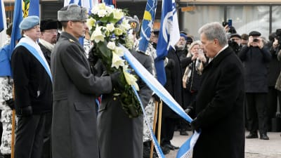 Sauli Niinistö håller i en blåvit banderoll som är fäst vid en blomkrans, omringad av människor, på Kaserntorget i Helsingfors.