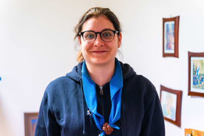 En ung kvinna med glasögon, mörkt hår i knut, blå luvtröja och scouthalsduk ser in i kameran och ler.