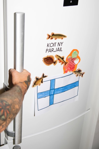 Jääkaapin ovi, jonka kahvaan on tarttunut tatuoitu käsi. Ovessa on kalamagneetteja, lapsen piirtämä suomenlippu ja lappu, jossa teksti "Koit ny pärjäil".