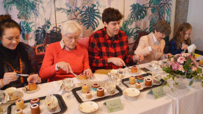Fem personer sitter på rad bakom ett bord och äter Runebergstårtor.