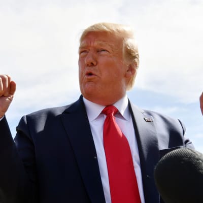 Donald Trump reagerar genom att knyta bägge händer. Han är fotad utomhus, iklädd mörkblå kostym, vit skjorta och röd slips. 