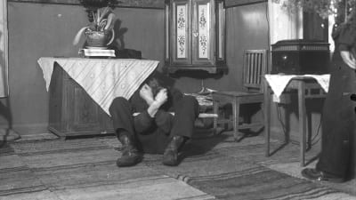 En man leker leken "lugg björn", och sitter på golvet med knäna uppåtdragna och armarna korslagda. Bilden är tagen 1938 i Närpes, Kalaks.