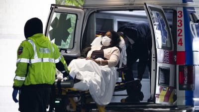 Kasvomaskiin puettu ja paareilla makaava nainen kannetaan ambulanssista sairaalan pihalle