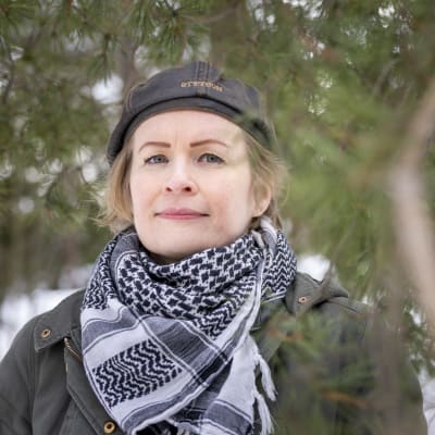 Hattuun ja huiviin pukeutunut nainen lumisessa metsässä.