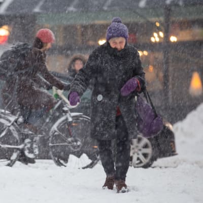 En äldre dam går över vägen i snöoväder, i bakgrunden en cyklist.