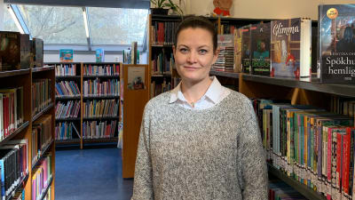 Kvinna i mörkt uppsatt hår och grå tröja står inne i ett bibliotek.