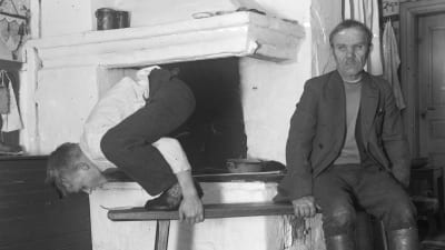 En man leker leken "ta sticka från långbänken", där han står på en bänk och hukar sig. Bilden är tagen 1936 i Strömfors, Kungsböle.