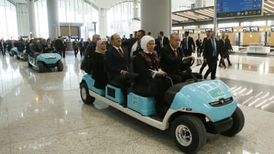 En bild på turkiets president Erdogan som åker i en liten taklös bil på en flygplats. Brevid honom sitter hans fru.