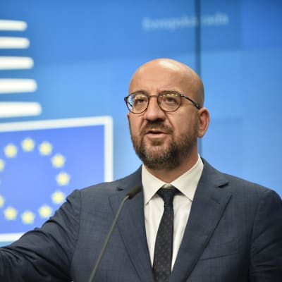 Charles Michel håller tal med handen utsträckt framför en blå bakgrund där EU:s flagga syns. Michel har runda glasögon, skägg och är skallig.