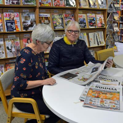 Två kvinnor sitter och läser en tidning vid ett bord i ett bibliotek.