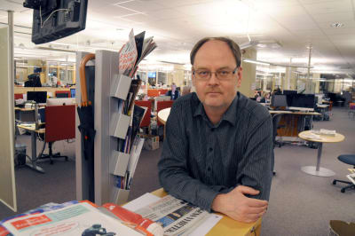 En man med glasögon står lutad mot ett bord. Bakom honom syns ett landskap från en tidningsredaktion.