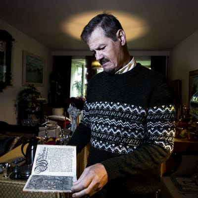 Veli Pekka Toropainen står i sitt vardagsrum och håller i en bok från 1600-talet.