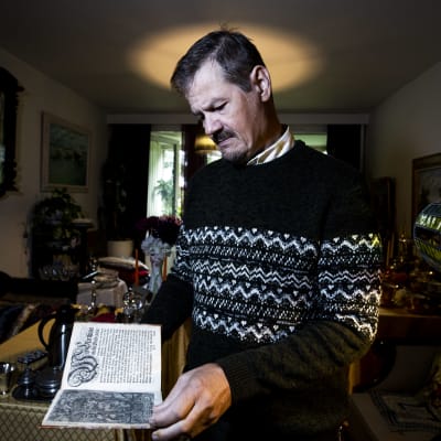 Veli Pekka Toropainen står i sitt vardagsrum och håller i en bok från 1600-talet.