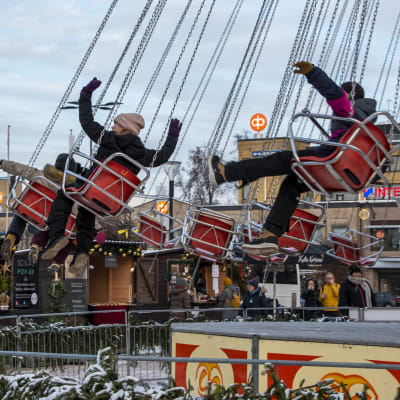 Barn åker i en karusell och viftar på armarna. I bakgrunden syns juldekorerade försäljningsbodar.