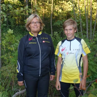 En kvinna och en pojke står bredvid varandra i en grön lummig skog. De ler och har på sig träningskläder. De har båda blont hår och kvinnan har glasögon.
