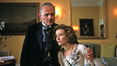 1900-luvun alun tyyliin herraskaisesti pukeutuneet mies (näyttelijä Anthony Hopkins) ja nainen (näyttelijä Emma Thompson) keltaisessa salongissa, nainen huolestuneen näköisenä nojaten mieheen, joka seisoo vakavana hänen rinnallaan ja pitää kättään naisen hartioiden ympärillä, kuva elokuvasta Talo jalavan varjossa