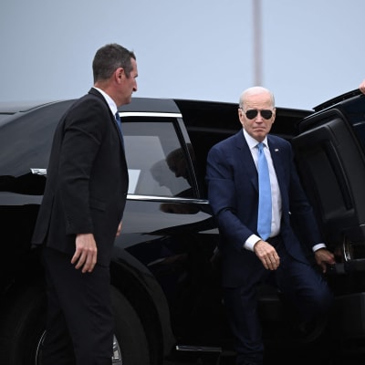 Joe Biden iklädd blå kostym och solglasögon stiger ut ur en svart bil imgiven av säkerhetsvakter.