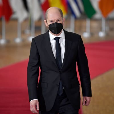 En man i mörk kostym och mörk slips (Olaf Scholz) med ett svart munskydd går längsmed en röd matta med EU.s flaggor uppradade i bakgrunden. 