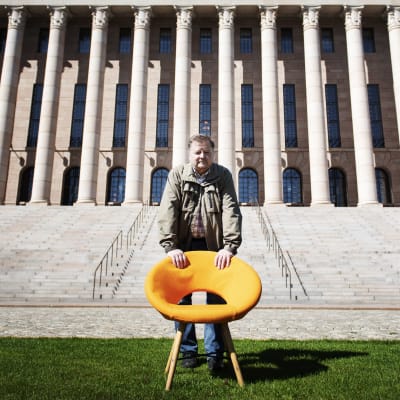 Mies seisoo suuren portaikon edessä nojaten oranssiin tuoliin.
