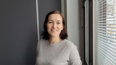 Kvinna med grå tröja står bredvid ett fönster.
