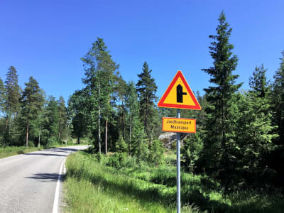 En asfalterad väg med skog på båda sidorna. Till höger syns ett trafikmärke, en triangel som varnar för en liten korsning och jordtransnporter.