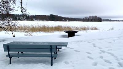 En blå bänk och ett blått bord står i snön vid en strand intill en grill.