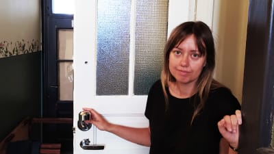 Mia Haglund har öppnat sin lägenhetsdörr och står och håller i den samtidigt som hon tittar in i kameran.