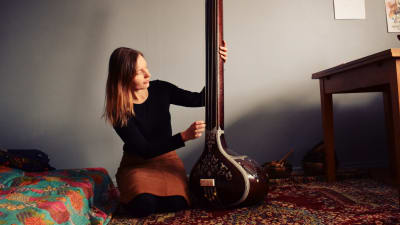 Laura Naukkarinen sitter på knä och spelar på ett indiskt stränginstrument i ett rum med turkisk matta och blå vägg.