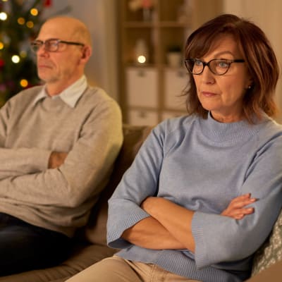 Medelålderns man och kvinna sitter i en soffa och ser sura ut, i bakgrunden en julgran