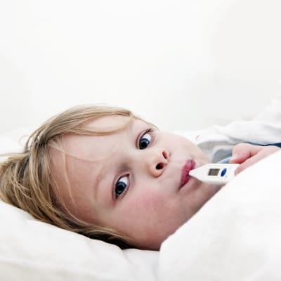 Ett barn ligger till sängs med feberröda kinder och en febertermometer i munnen.