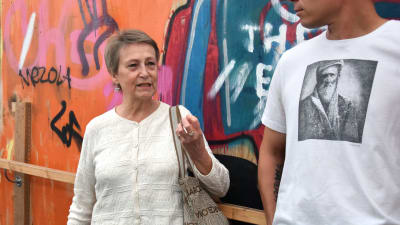 Kvinna i vit blus och med kort hår talar med en man i vit t-skjorta. I bakgrunden graffiti i olika färger.
