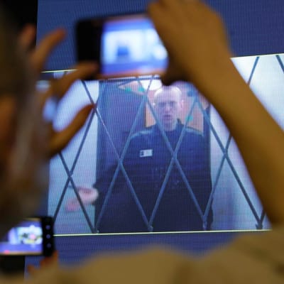 Aleksej Navalnyj bakom ett galler på en skärm. I förgrunden håller en kvinna upp en mobiltelefon för att ta en bild av skärmen som visar Navalnyj.
