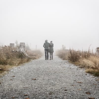 Två personer håller om varandra och går längs en grå väg. Bilden är tagen bakifrån.