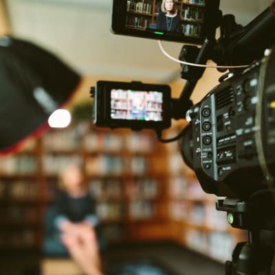 En tv-intervju pågår, en kvinna intervjuas i ett bibliotek, filmkameran är i fokus och en lampa som lyser mot kvinnan syns också i förgrunden.