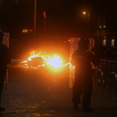 Kravallpolis vaktar ett brinnande fordon på en i övrigt mörk gata.