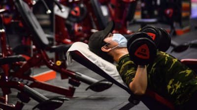 En man i munskydd lyfter vikter på ett gym.