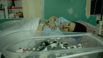 En kvinna ligger i en sjukhussäng. Hon är vänd åt sidan och tittar på en bebis som ligger bredvid.